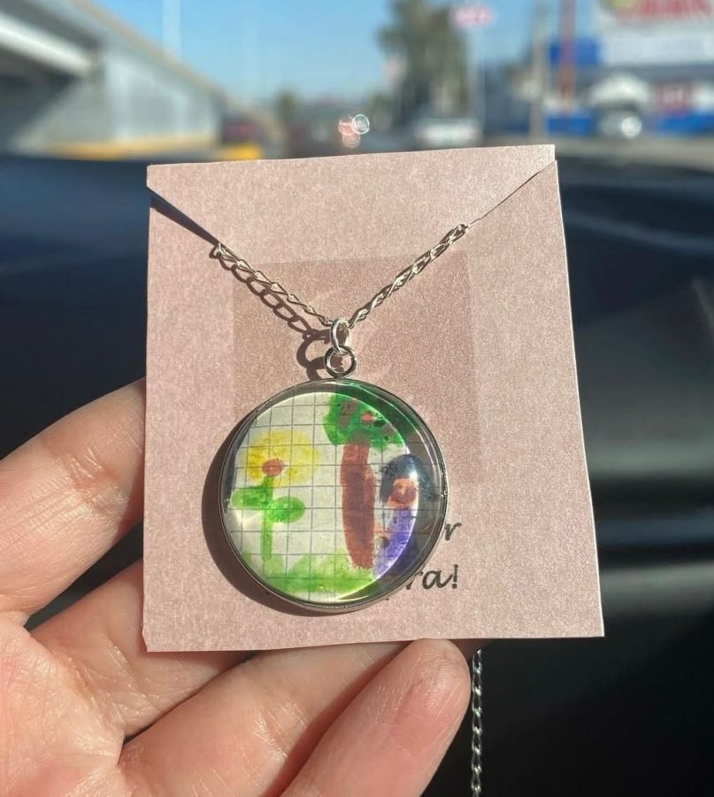 Collar con un paisaje dibujado a lápiz de color, presentado en una tarjeta de papel kraft.